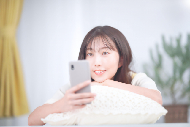 クッションに持たれながらスマホを見ている女性の画像