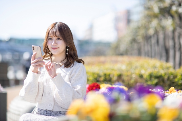 花壇の前でスマホを持ち笑顔でほほ笑む女性の画像