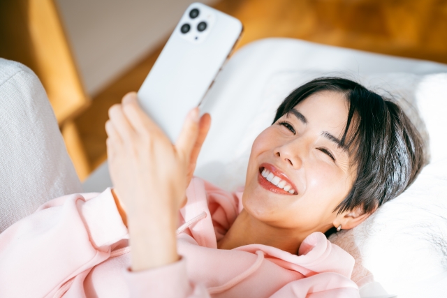 ベッドに寝そべってスマホでスマホを見ている女性の画像