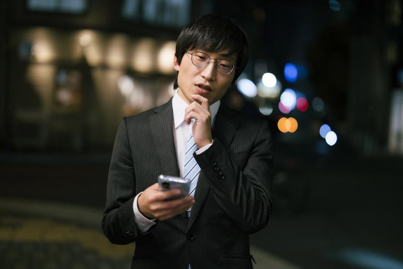 夜道でスマホを操作しながら悩んでいる表情の男性の画像