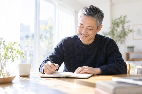 明るい窓辺のテーブルで、手帳にこれからの進め方を書き出している40代後半の男性