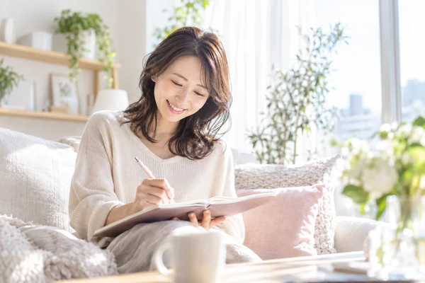 明るい窓辺のソファで、手帳に婚活にかける予算を書き込んでいる30代の女性