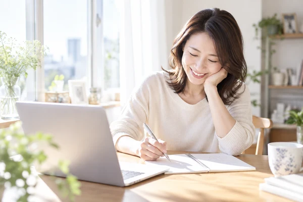 自宅の窓辺の机で、結婚相談所の料金をノートに書き出して見比べている30代の女性
