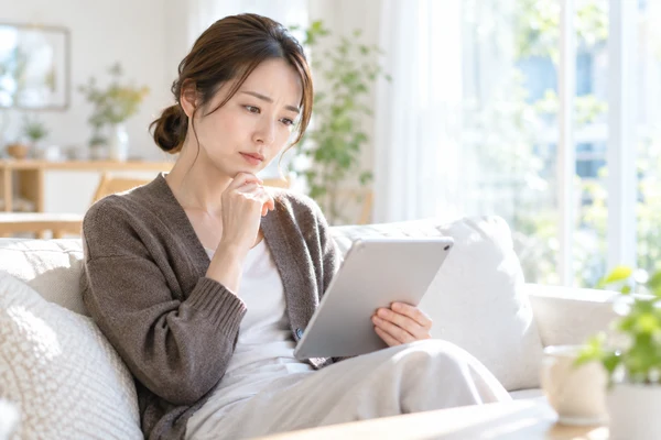 明るいリビングのソファでタブレットを持ち、結婚相談所のページを見ている30代前半の女性
