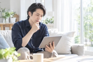 明るいリビングのソファでタブレットを持ち、これからの進め方を考えている30代後半の男性