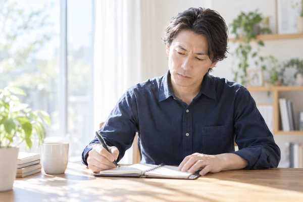 自宅の窓辺の机で手帳を開き、この3カ月の活動の数を書き出している30代後半の男性