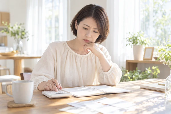自宅の窓辺の机で電卓とノートを広げ、1年目にかかる費用を計算している40代前半の女性