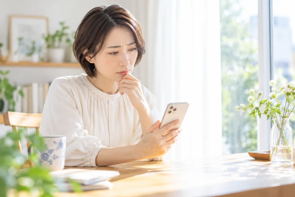 自宅の窓辺の机でスマートフォンを持ち、担当者とのやり取りを見返している40代前半の女性